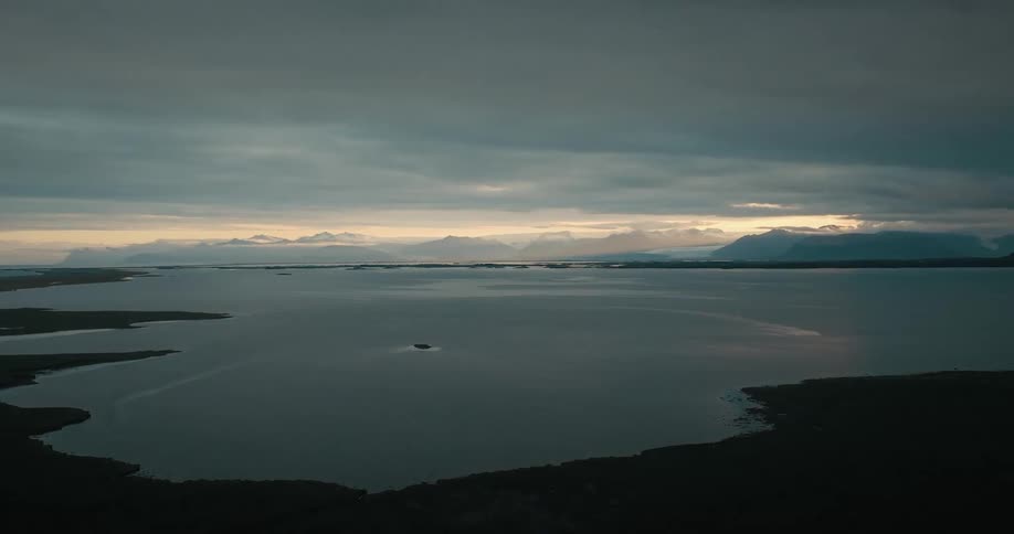 A moody and serene wide-angle shot of a vast fjord at dusk, with distant mountains under a dramatic, cloudy sky