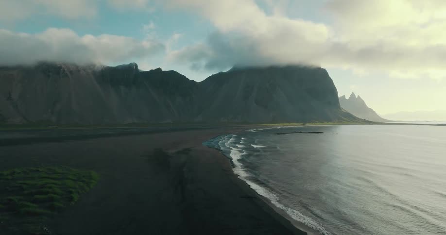 Gentle ocean waves wash ashore on a black sand beach with a majestic, cloud-covered mountain range in the background