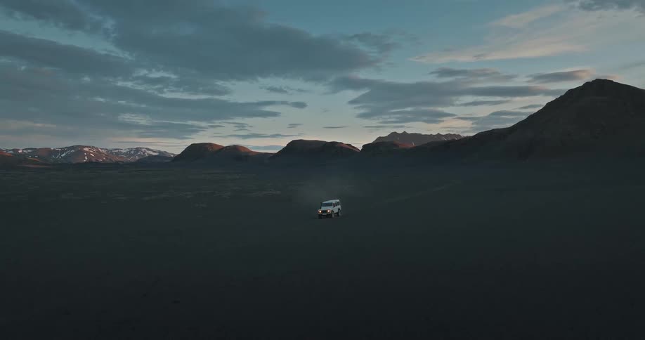An aerial view shows a lone white SUV driving through a vast, remote volcanic desert in Iceland at dusk