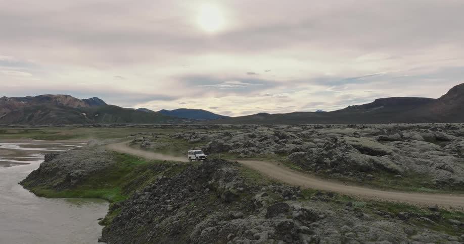 An aerial shot follows a white off-road vehicle as it drives through the vast, moss-covered volcanic landscape of the Icelandic highlands