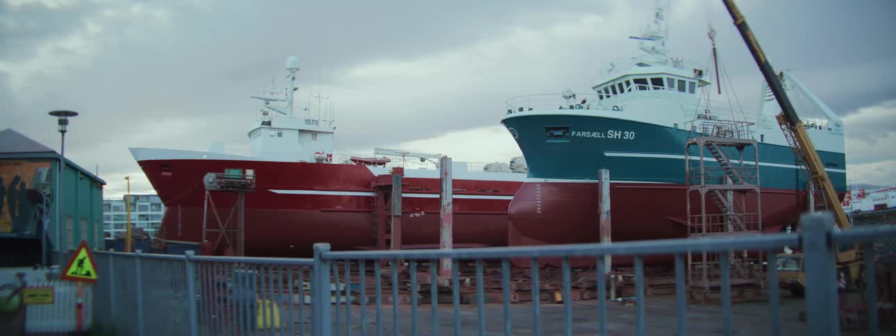 A pair of large fishing trawlers rest in a dry dock at a shipyard under an overcast sky