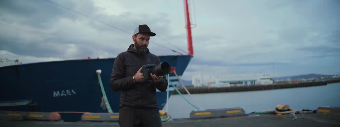 A bearded photographer in a baseball cap raises his camera to take a picture while standing on a dock in an industrial harbor