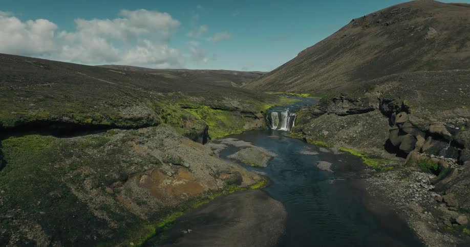 An aerial view flies over a wide river valley towards a beautiful hidden waterfall in the dramatic highlands of Iceland
