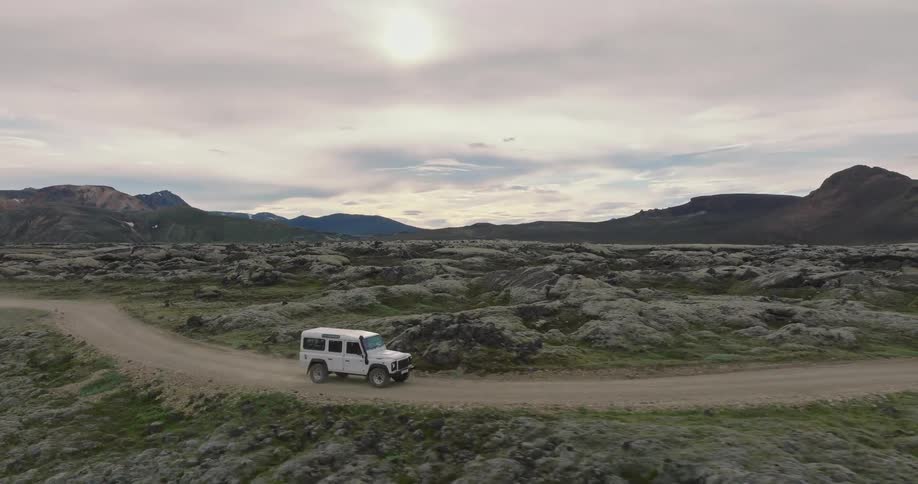 Aerial view of a white off-road vehicle driving along a dusty dirt track through a vast, moss-covered volcanic lava field in Iceland