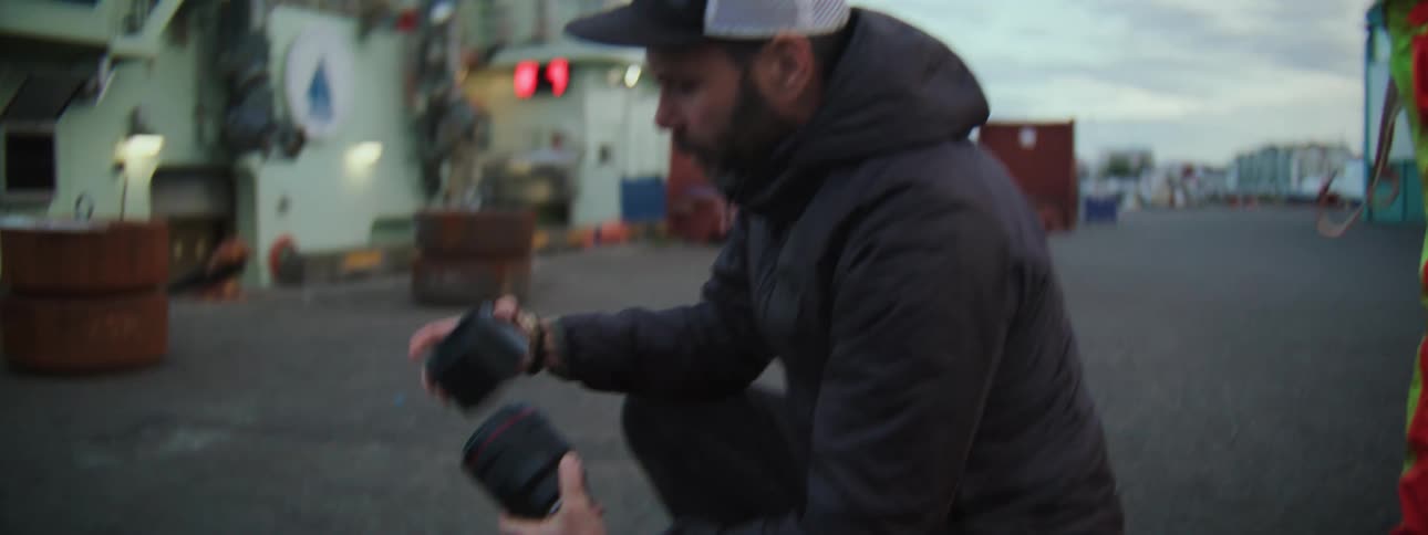 A professional photographer wearing a cap kneels down on a dock to prepare his camera equipment for a shoot