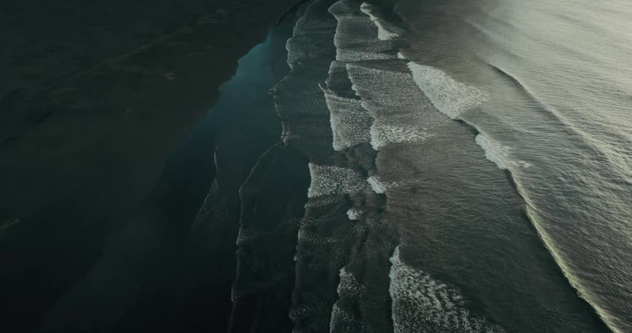 Aerial drone view slowly tilts down over a moody black sand beach with ocean waves rolling in at the base of dramatic mountains