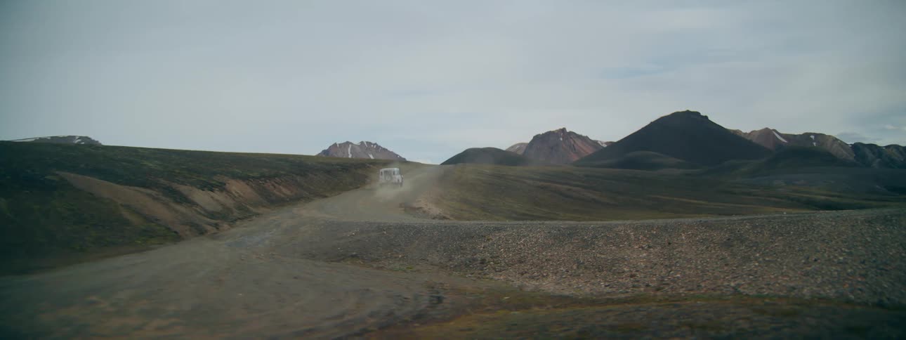 A white off-road vehicle drives down a winding dirt road, kicking up dust in a vast, remote mountain landscape
