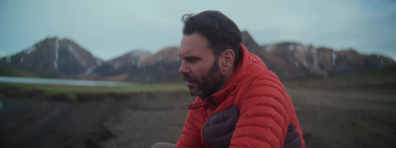 A pensive man in a red jacket takes in the view of a remote, mountainous landscape