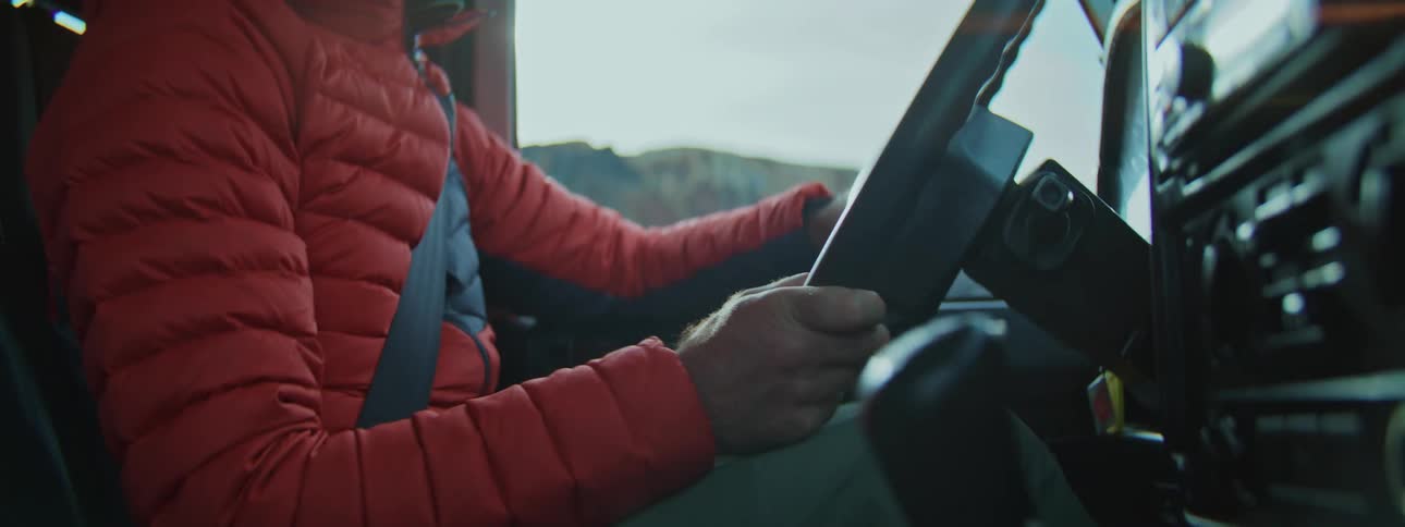 An adventurer wearing a red jacket grips the steering wheel and shifts gears while driving an off-road vehicle through the mountains