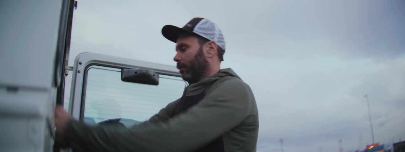 A man in a baseball cap layers up with a hoodie and a warm puffer jacket by his van in slow motion