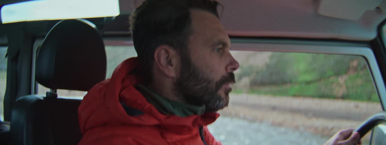 A focused, bearded man in a bright red jacket drives a vehicle through a rugged, green landscape