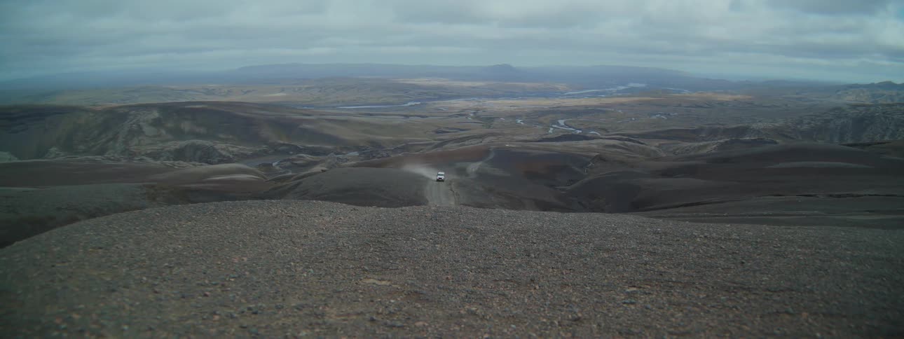 A lone white SUV drives down a winding dirt road through a vast and desolate Icelandic valley