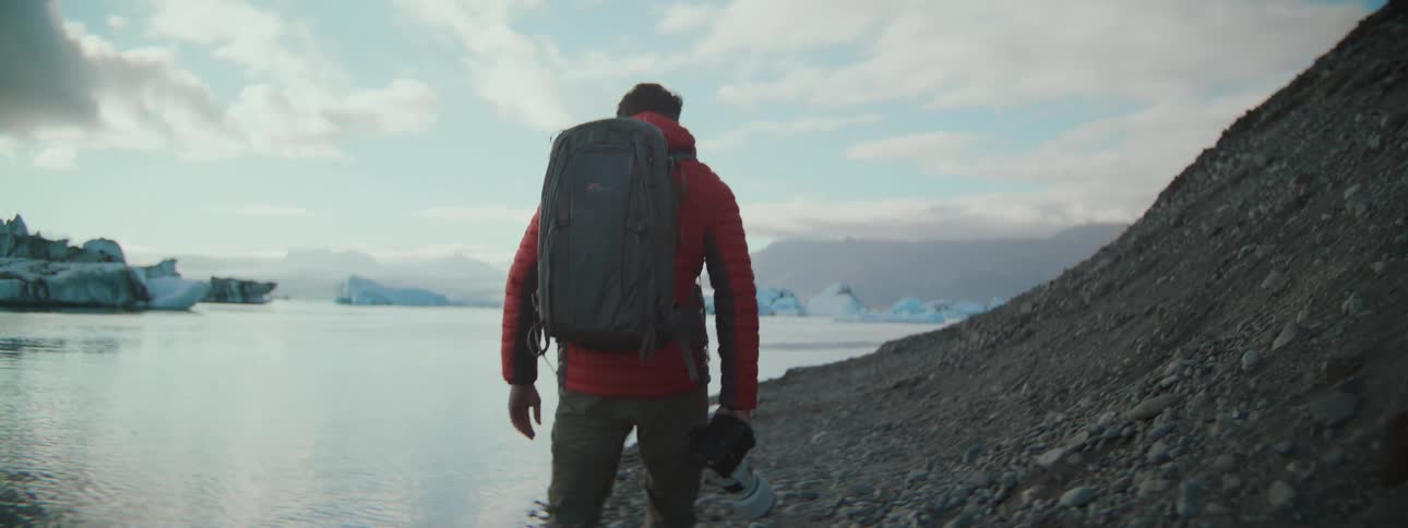 A photographer with a backpack and camera walks along the rocky shore of a glacial lagoon with icebergs