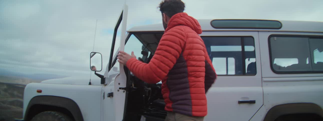 A bearded man in a red puffer jacket gets out of his white truck in a remote, scenic landscape in slow motion