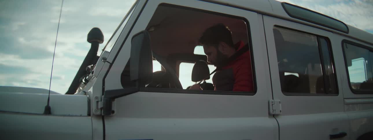 A bearded man in a red jacket buckles his seatbelt, preparing to drive his white off-road vehicle on an adventure