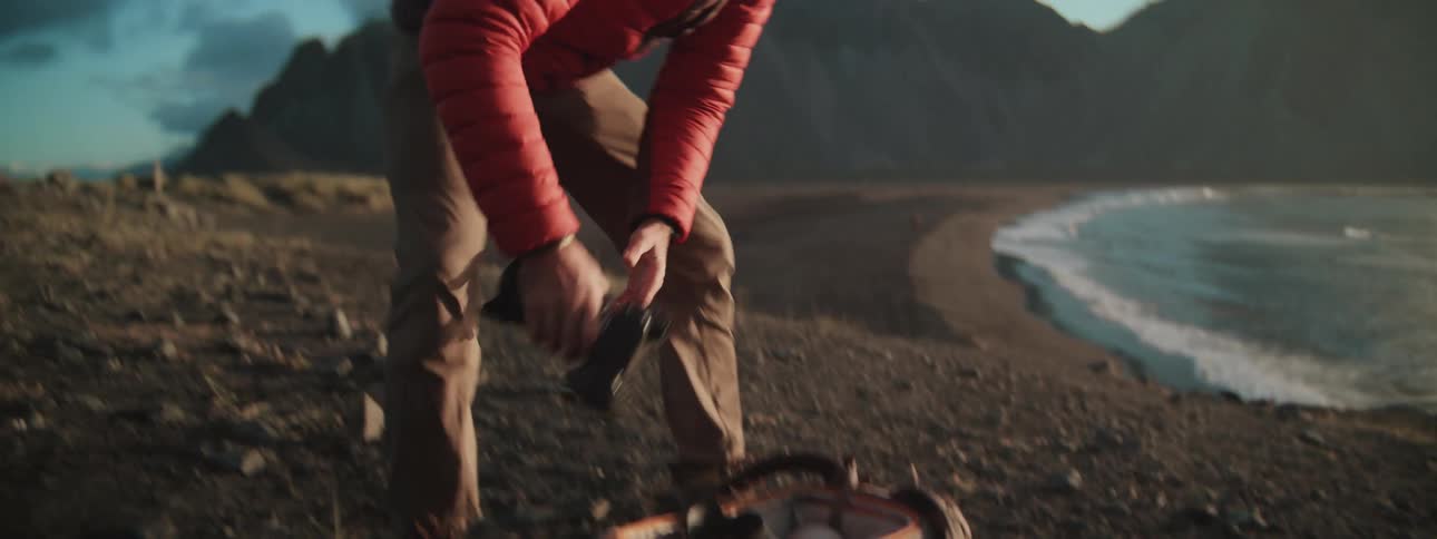 An adventurous male photographer assembles his camera gear while standing on a cliff overlooking a scenic beach and mountains