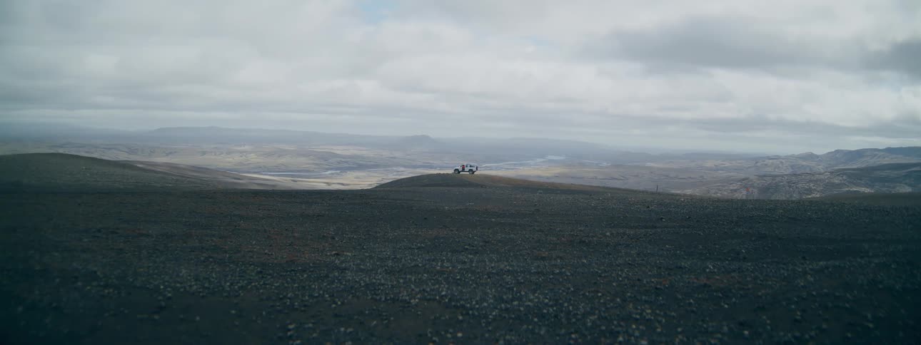 A white SUV is parked on a vast, barren volcanic plateau as travelers explore the epic, remote landscape