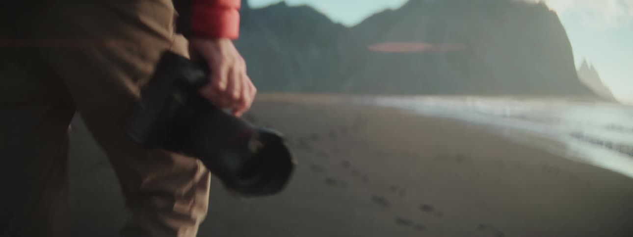 A photographer in a puffer jacket walks along a remote, sandy beach with a camera in hand during the golden hour