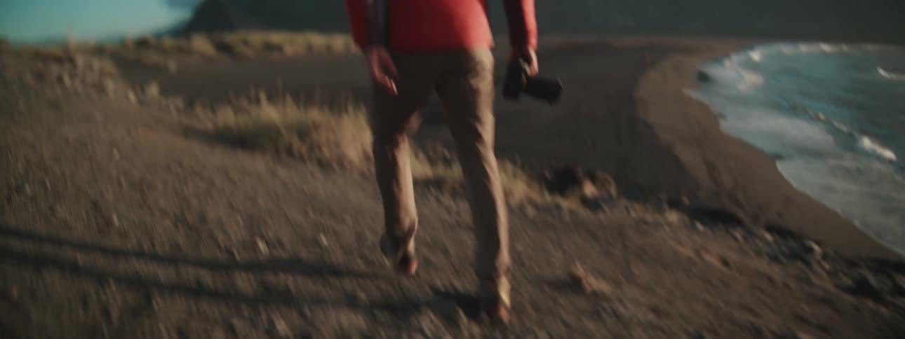 A male photographer in a red jacket walks with his camera down a dark, sandy hill toward a beautiful mountain range and the ocean