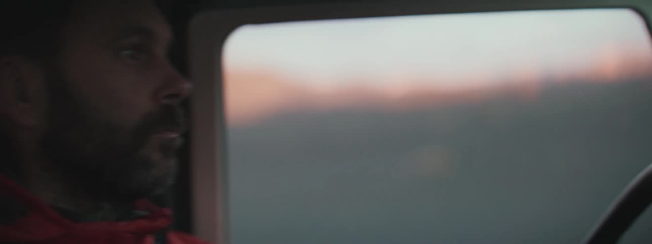 A close-up shot of a bearded man driving his car at sunset, with the camera panning from his face to his hand on the steering wheel