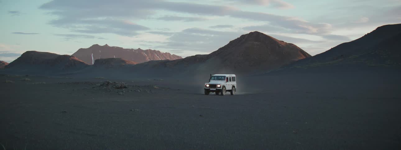 A white off-road vehicle drives across a desolate, dark, sandy plain with mountains in the background at dusk