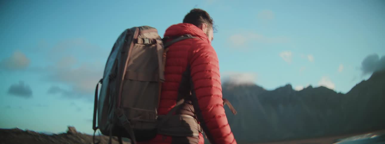 A photographer wearing a red jacket and a backpack hikes with his camera through a rugged mountain landscape towards a coastal beach in slow motion