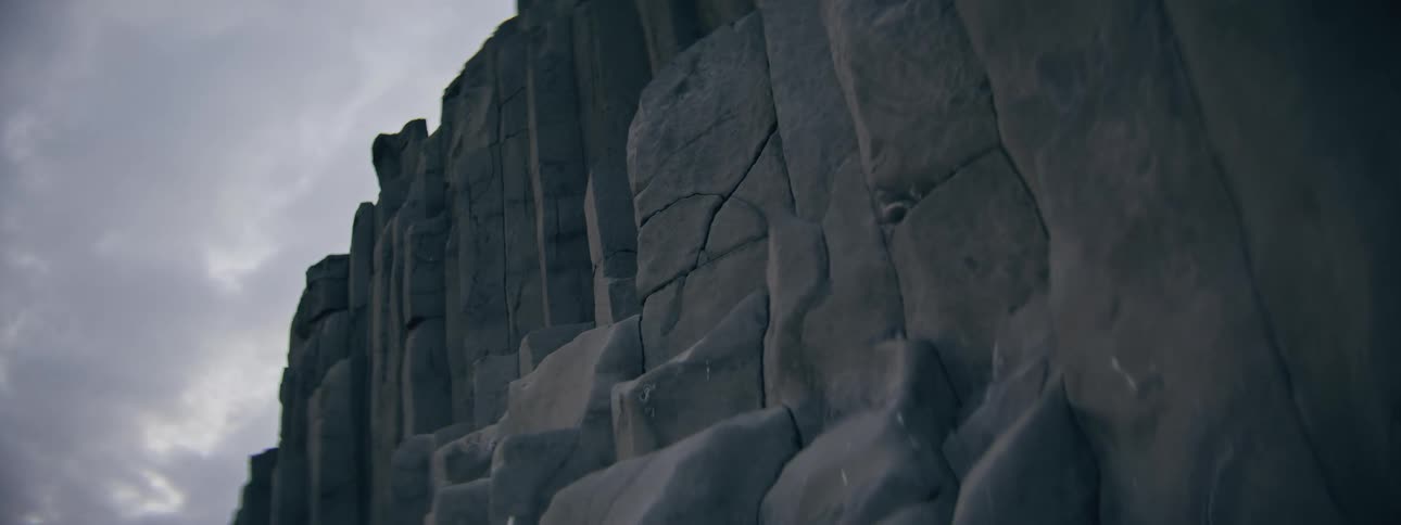 Raindrops fall on a dark, dramatic basalt rock formation under a grey sky