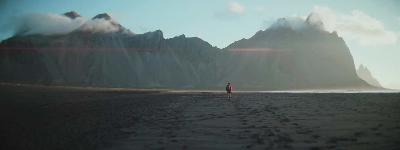 Cinematic wide shot of a lone hiker walking across a desolate black sand beach towards cloud-covered mountains