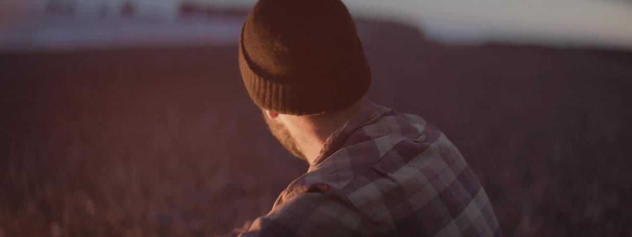 A bearded man in a beanie and plaid shirt tends to a warm campfire at dusk