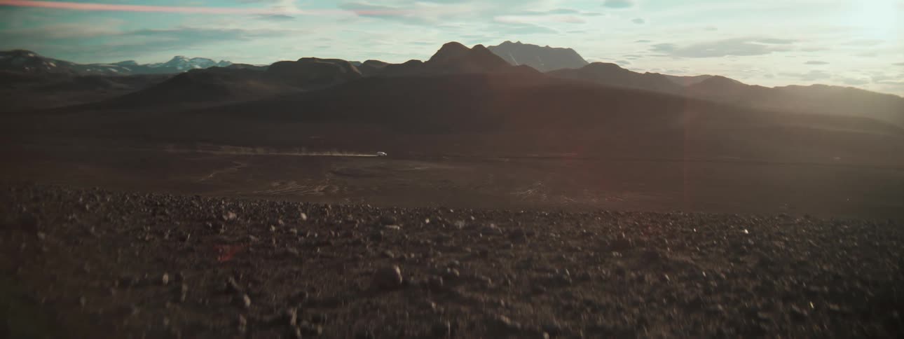 A lone white vehicle kicks up dust while driving through a vast, desolate rocky landscape with mountains in the background at sunset