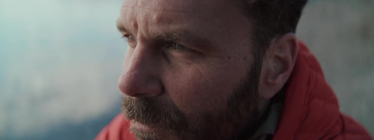 A close-up shot of a pensive, bearded man in a red jacket, sweating as he thoughtfully gazes into the distance