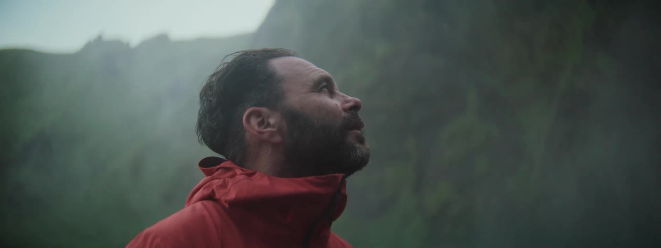 Slow motion close-up of a man in a red jacket looking up in awe at a misty mountain landscape