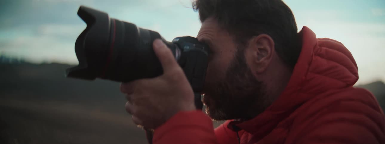 A male photographer in a red jacket focuses through his professional camera's viewfinder to capture the outdoor landscape during golden hour