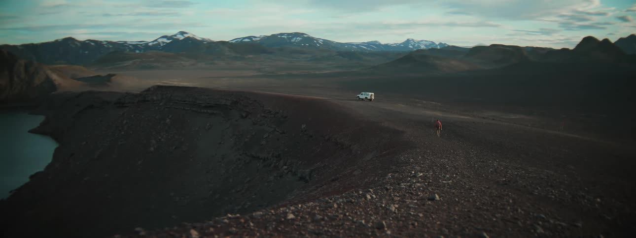 A lone explorer hikes along the rim of a volcanic crater in a vast and remote landscape with an off-road vehicle parked in the distance