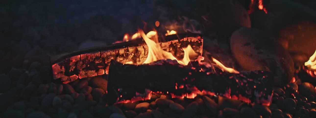 A mesmerizing slow motion close-up of a campfire with burning logs and glowing embers on a bed of rocks at night