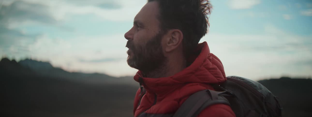 A male hiker in a red jacket looks out at the scenic mountain view as the sun flares behind him