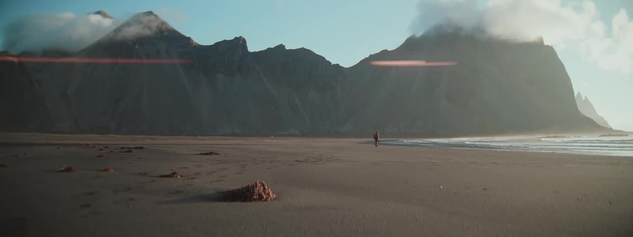 A solitary adventurer walks along a vast, desolate beach, dwarfed by an epic mountain range