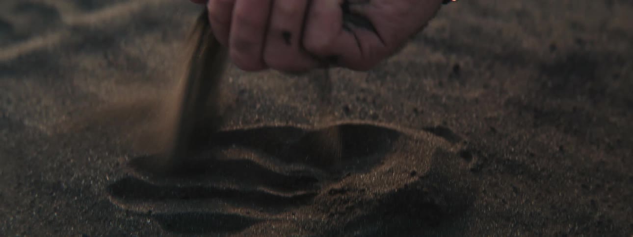 A close-up shot of a man's hand drawing lines in dark volcanic sand before letting it slip through his fingers in slow motion