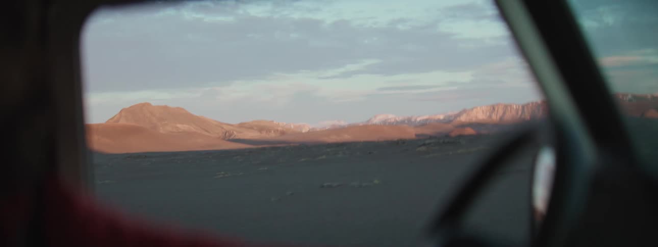 Point-of-view from a moving car captures the vast, desolate landscape and distant snow-capped mountains at sunrise
