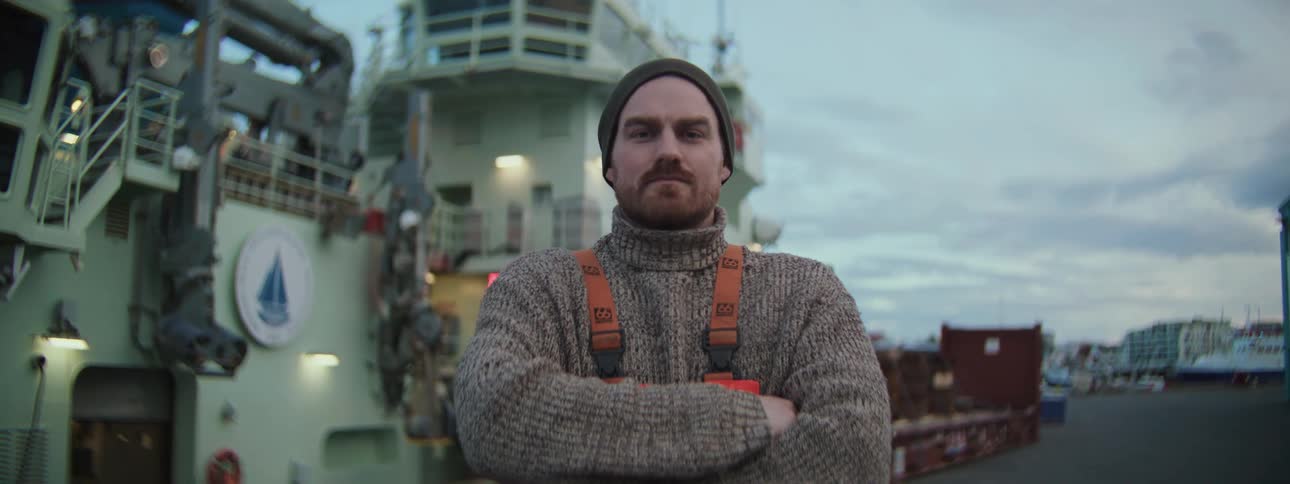 A confident fisherman in a wool sweater and beanie stands with arms crossed in front of his fishing boat at the dock as the camera slowly pushes in