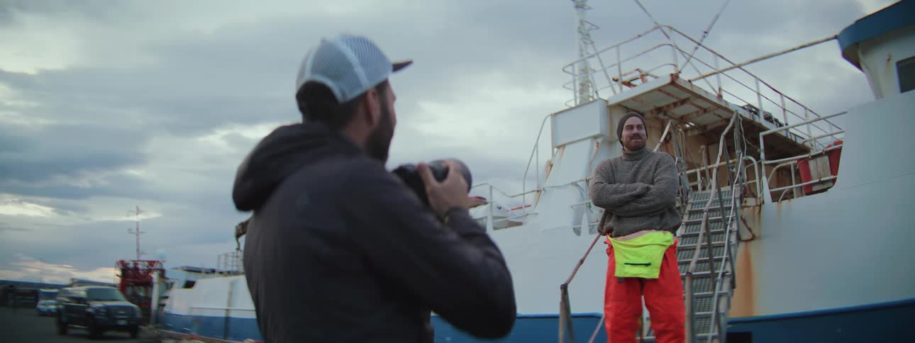 A photographer takes a portrait of a rugged fisherman posing in front of a large fishing trawler at the docks