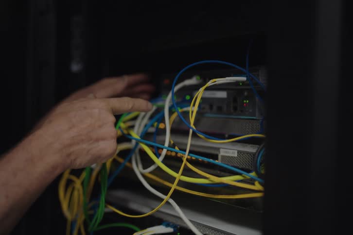 An IT technician's hands manage and point to tangled network cables connected to a server rack in a dark data center