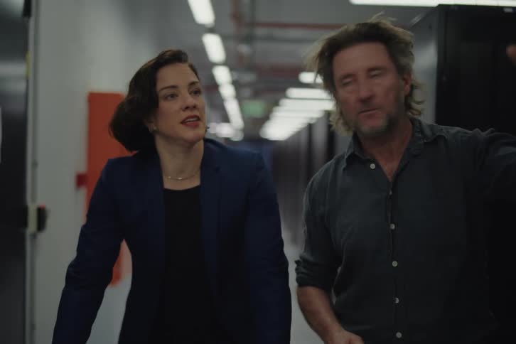 An IT technician explains a critical system issue to a female manager as they walk through a data center server room
