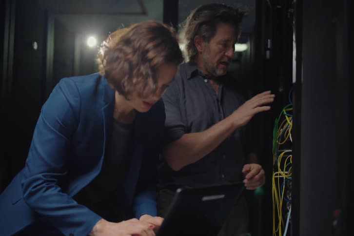 Two IT specialists collaborate on troubleshooting a network issue inside a dimly lit server room, with one typing on a laptop and the other inspecting the server rack