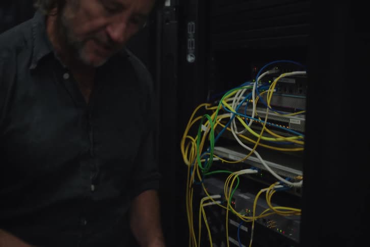 An IT technician explains a server rack's complex wiring to a colleague using a laptop for diagnostics in a dark data center