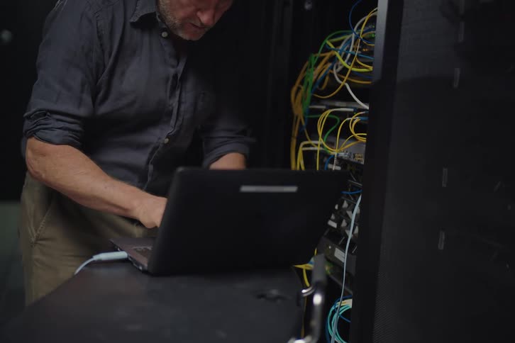 A focused IT technician with windblown hair works on network cables in a server rack using a laptop in a data center