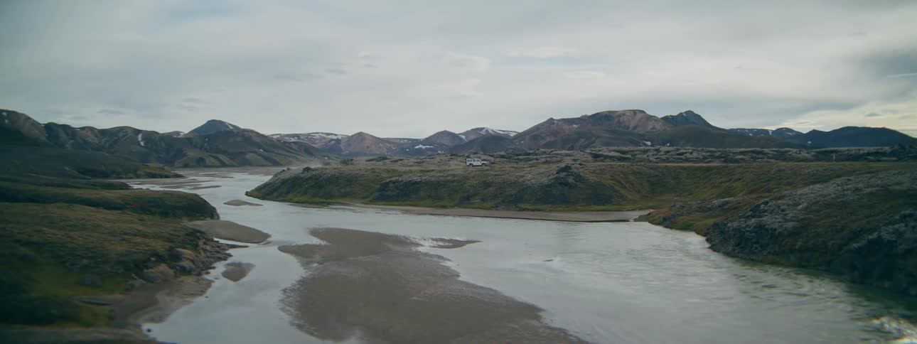 An off-road vehicle drives along a rugged dirt path next to a wide river in the vast Icelandic highlands