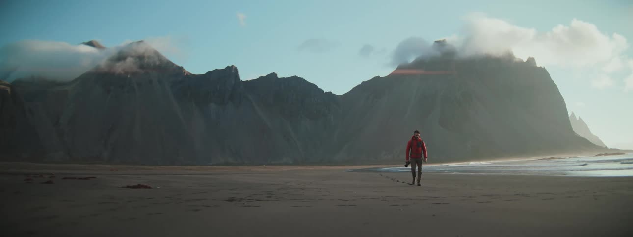 Slow motion shot of a man in a red jacket walking on a vast, sandy coastline with stunning mountains in the background