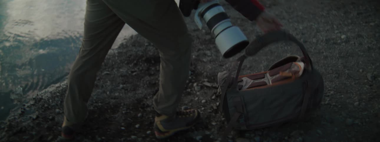 A photographer in a red jacket packs away his camera equipment into his backpack on the rocky shore of an icy glacial lagoon
