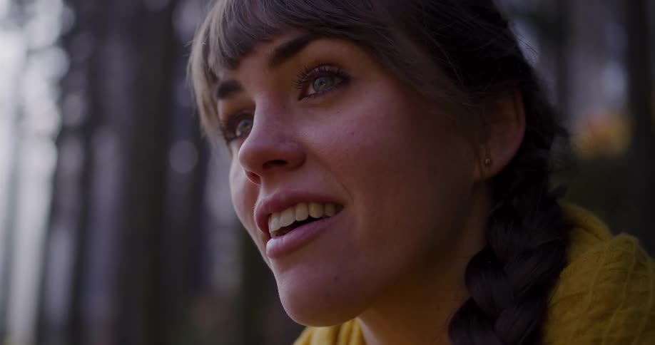 A close up of a thoughtful young woman with a braid looking up with a serene expression while surrounded by a tranquil forest